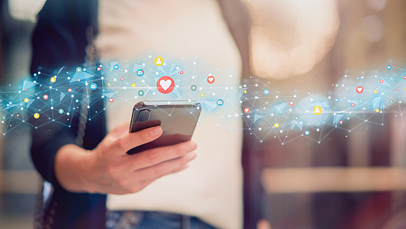 © Sitthiphong – istock.com Eine Person hat ein Handy in der Hand und um das Handy herum sind Social Media Icons zu sehen
